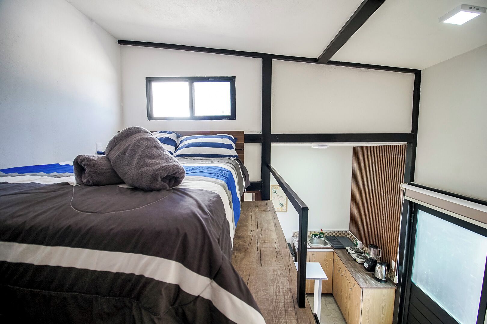 Quarto com cama, janela, uma mesinha e um espaço com cozinha compacta.