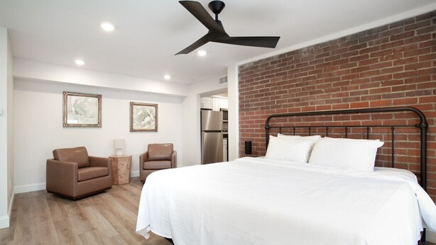 Un dormitorio moderno con una cama grande, ventilador de techo, pared de ladrillo decorativa y una kitchenette al fondo.