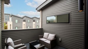 Een balkon met rieten meubels en een glazen tafel, met uitzicht op een rij moderne gebouwen.