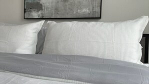Una cama bien hecha con sábanas blancas y una colcha texturizada, un cuadro abstracto en blanco y negro en la pared y un cabecero sencillo y moderno.