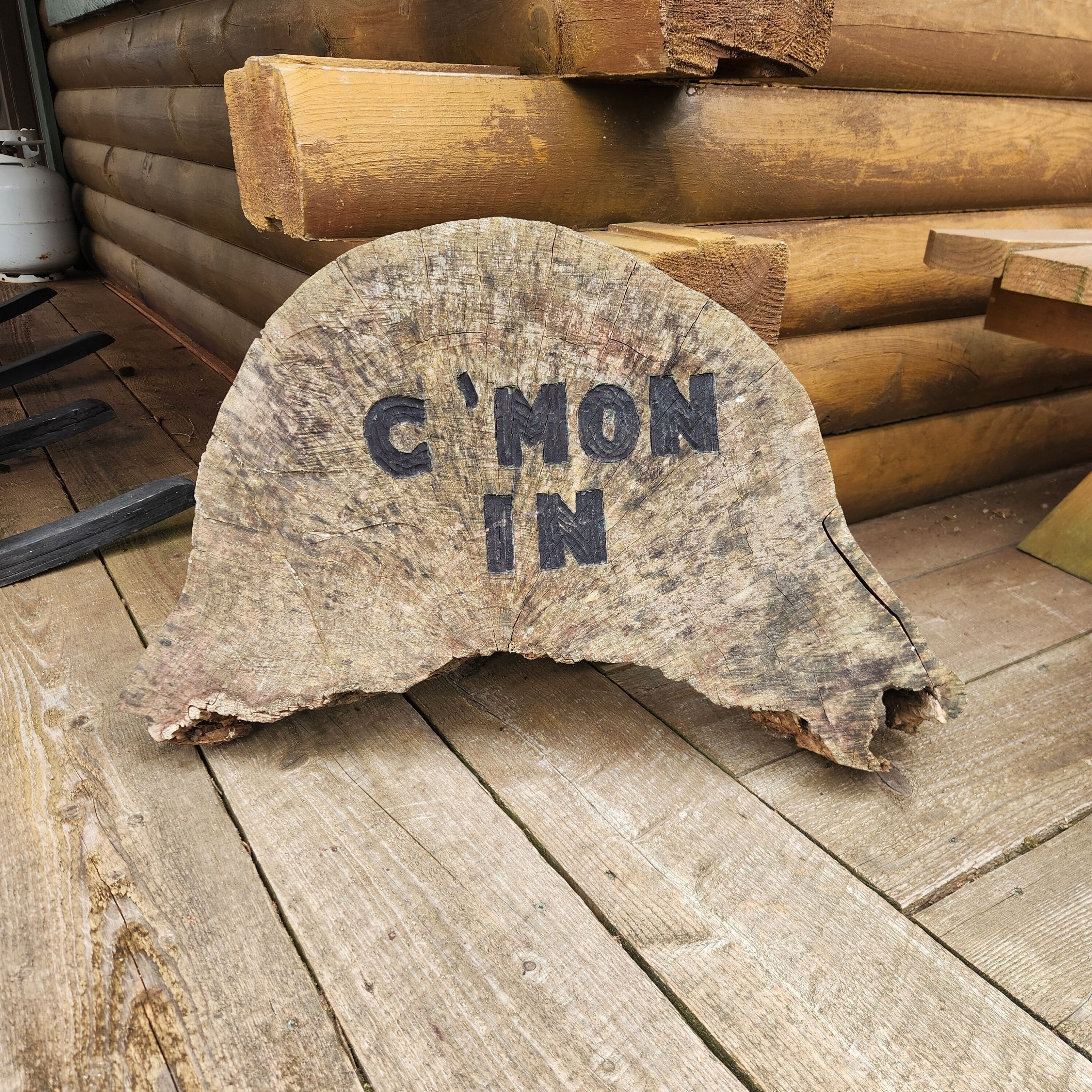 Un objet rustique en bois, sur lequel est gravé le texte « C’NON IN », posé sur un plancher en bois.