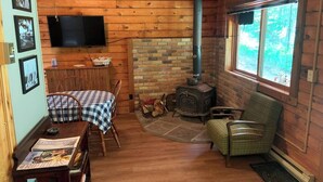 A cozy cabin interior with a brick fireplace, a television mounted on the wall, a dining table with chairs, and a window with a view of the outdoors.