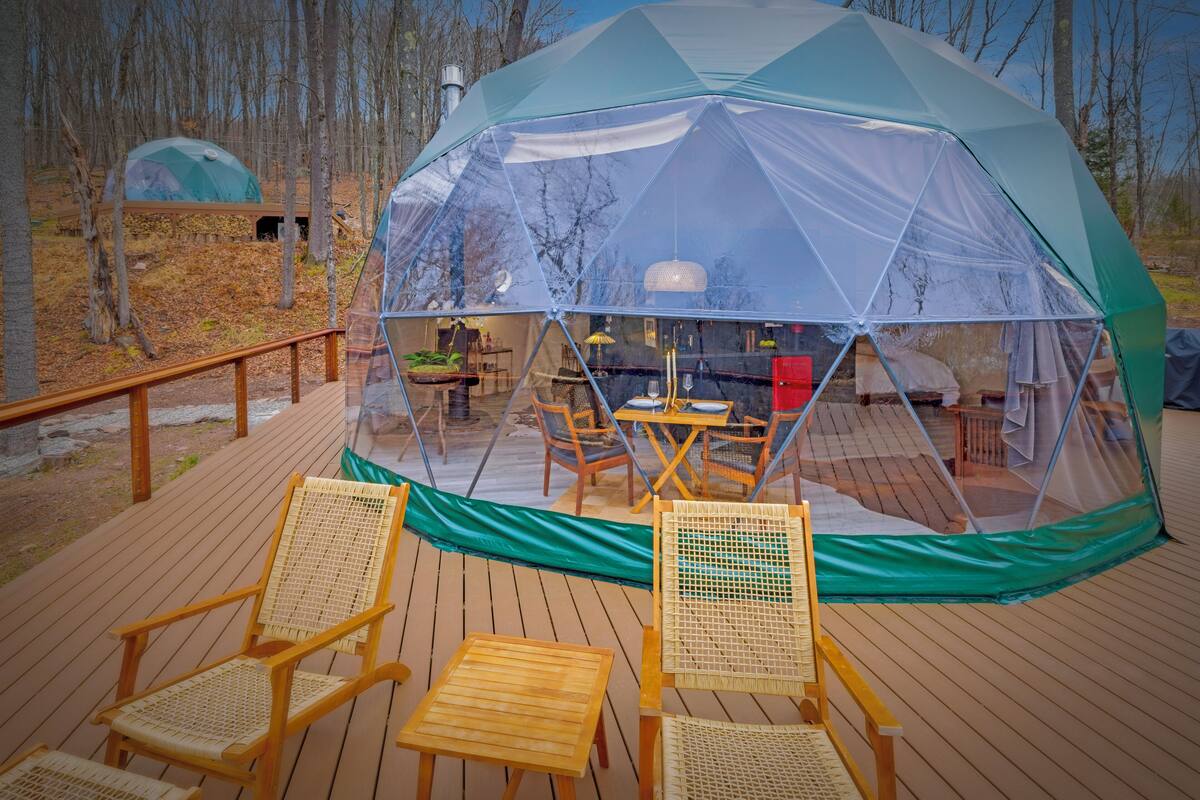 A transparent geodesic dome tent with a wooden deck, two wicker chairs, and a dining table with chairs.