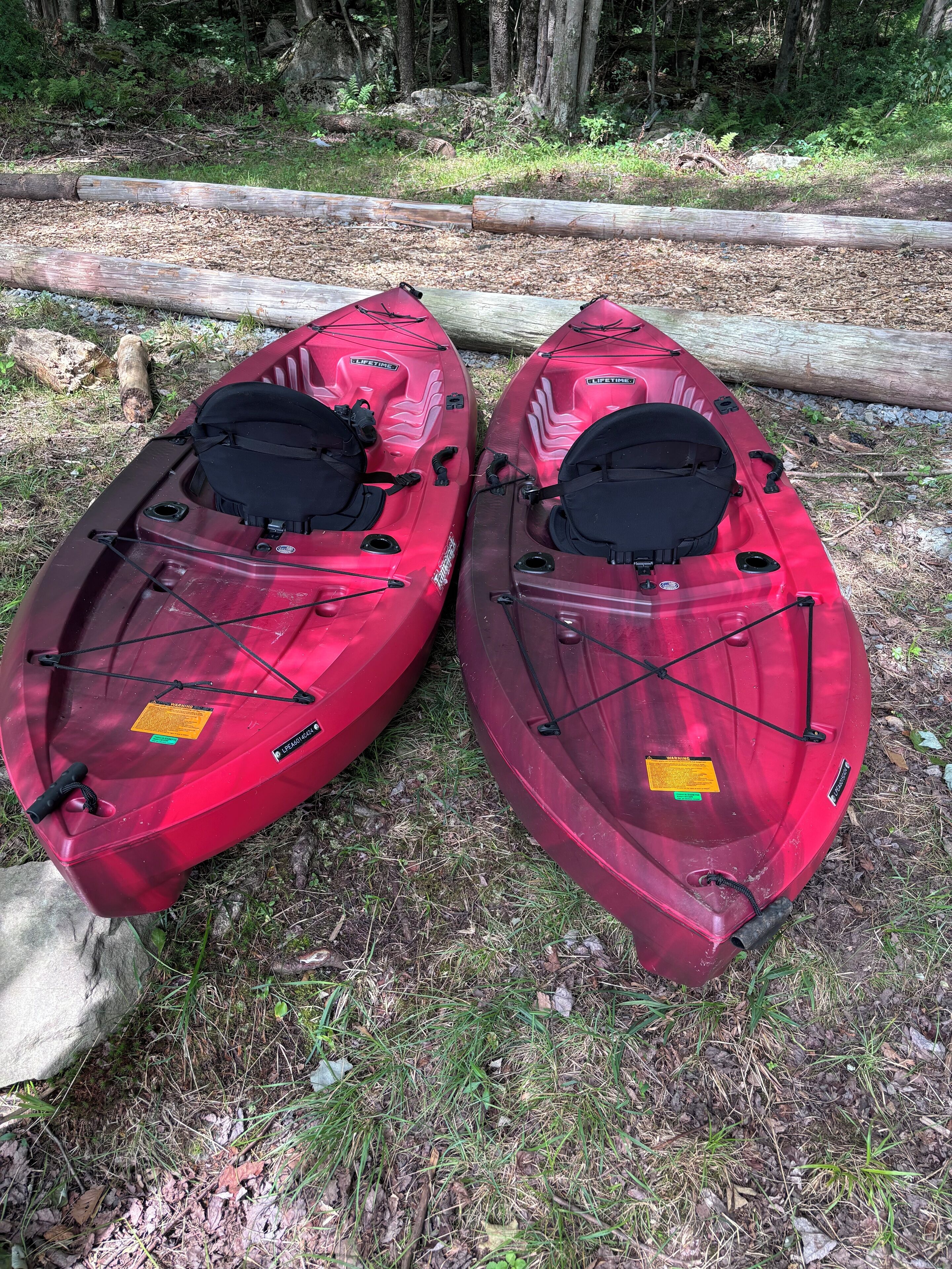 Dos kayaks rojos con asientos y chalecos salvavidas negros, ubicados en una zona de césped cerca de un tronco.