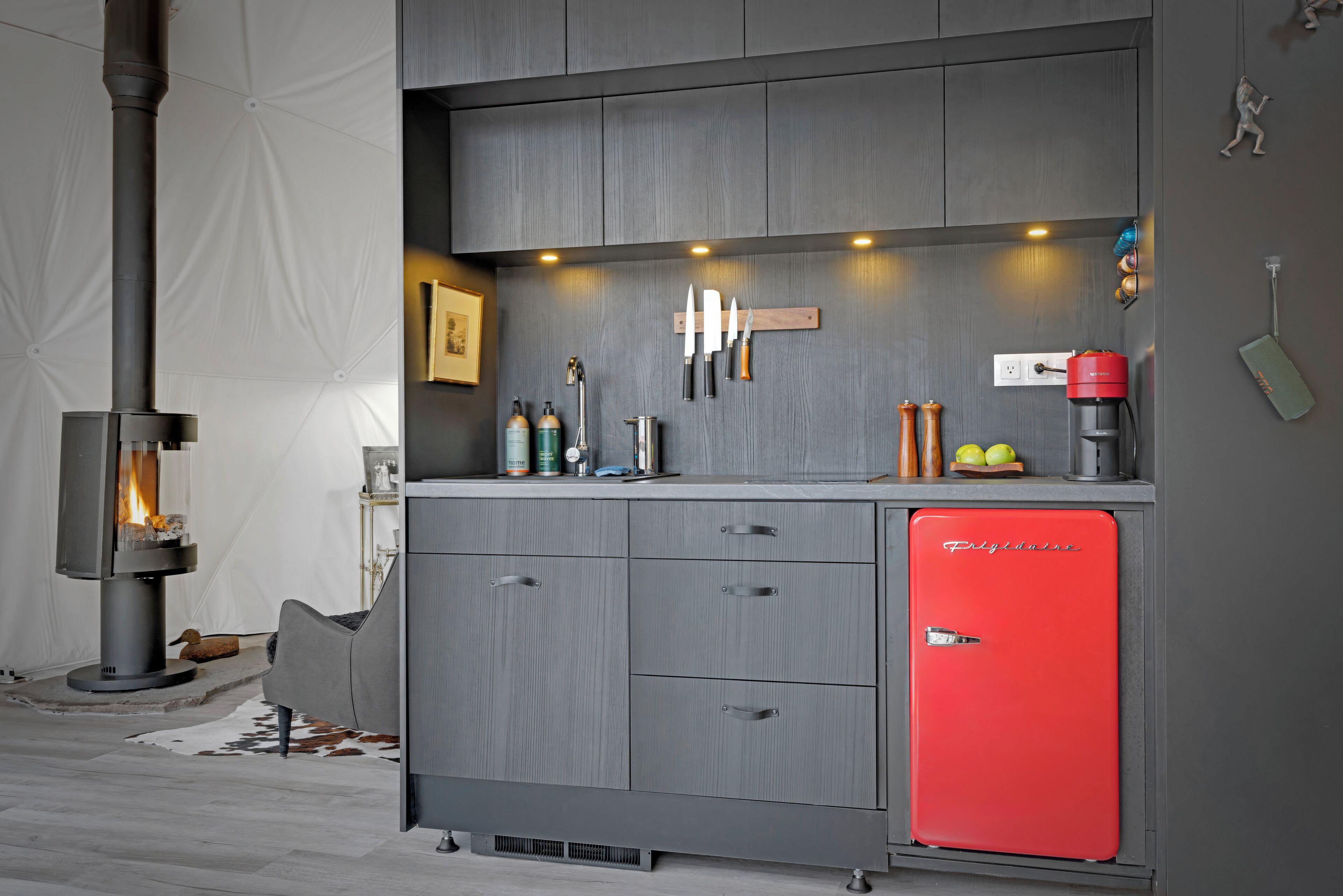 A modern kitchen with grey cabinetry, a red refrigerator, and a wood-burning stove.