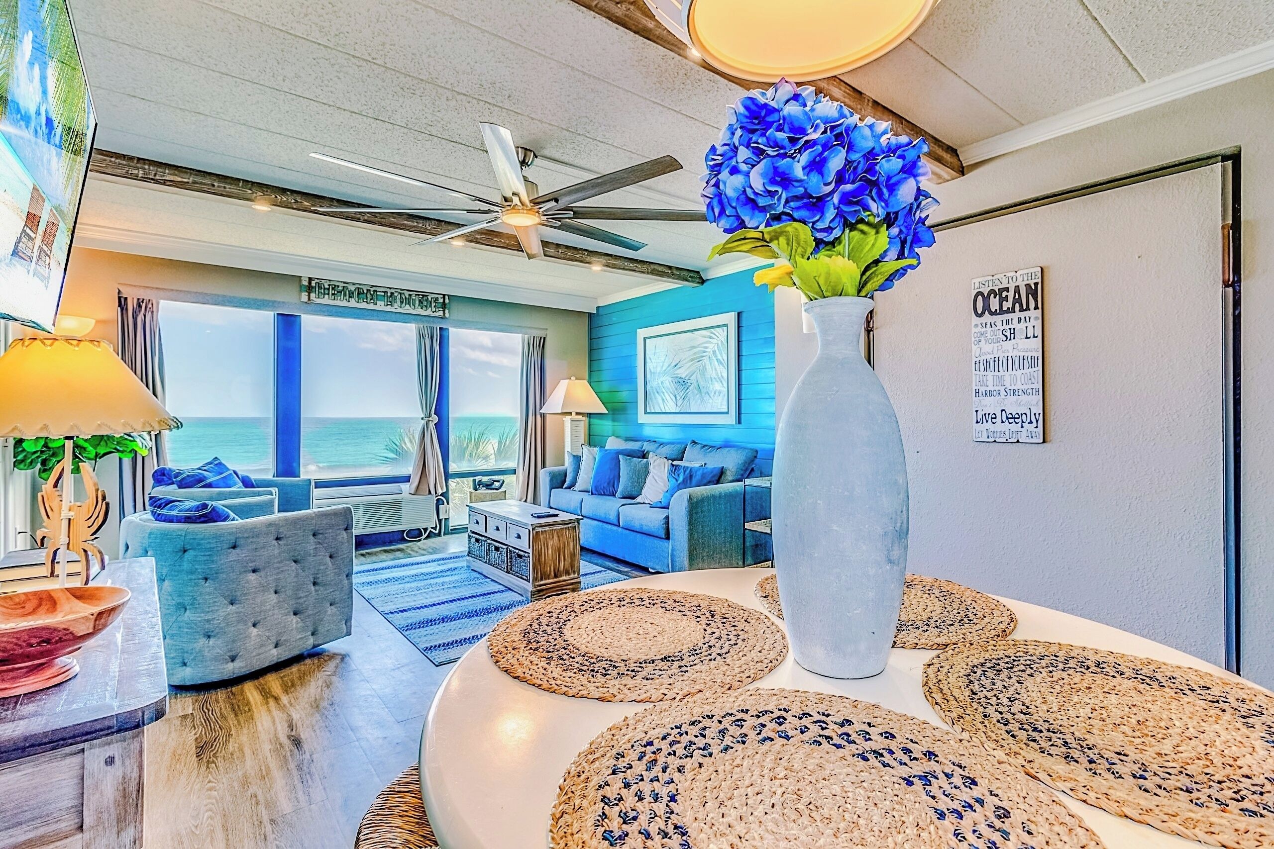 Un soggiorno con vista sull'oceano, una ventola a soffitto, un vaso grande con fiori blu e un cartello appeso al muro.