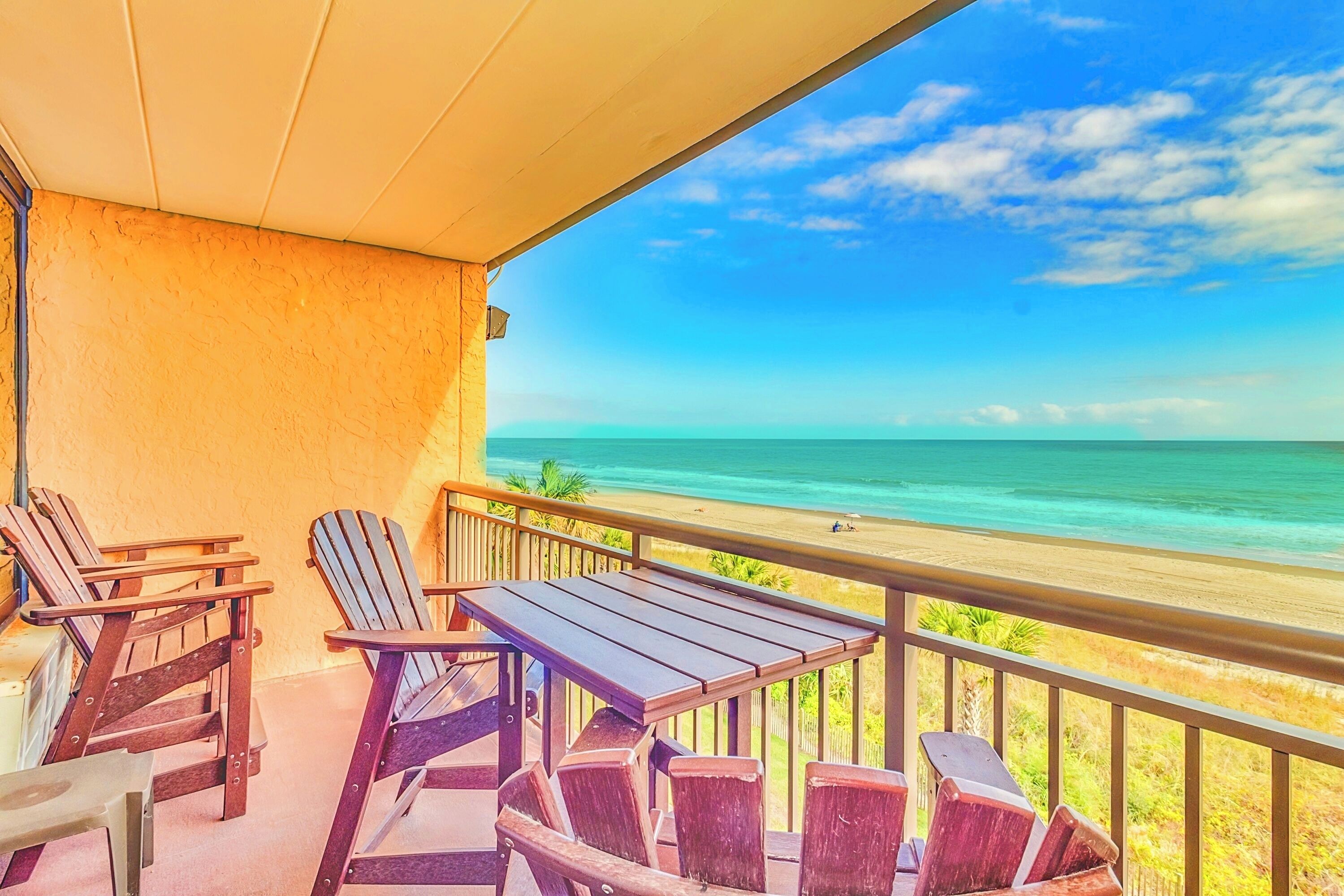 A balcony with wooden chairs and a table overlooking a beach.