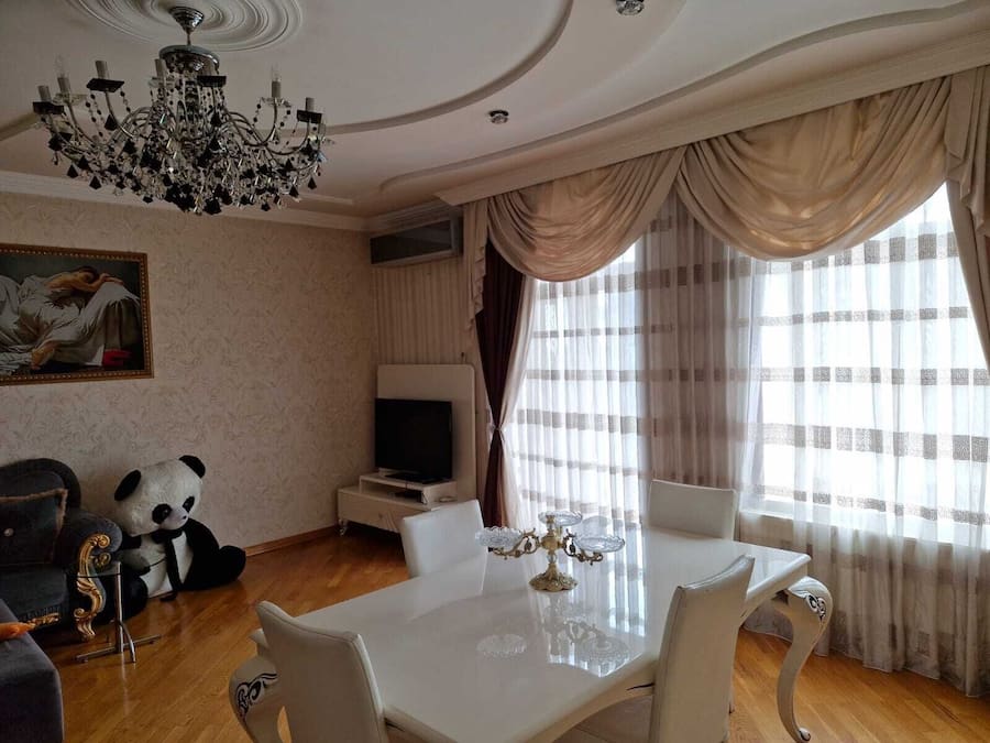 Uma sala de estar ampla, com uma mesa de jantar branca, assentos confortáveis, um lustre grande e uma televisão.