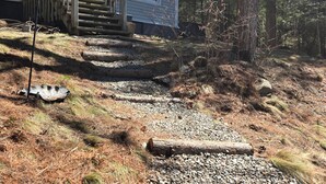A house with a wooden staircase leading to a porch, surrounded by trees and a gravel path.