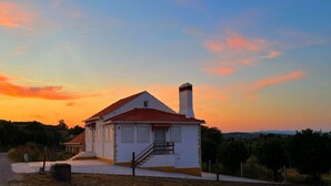 A white building with a red-tiled roof and a chimney, surrounded by greenery and a clear sky.