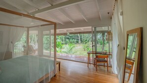 A bedroom with a four-poster bed, a wooden desk, and a chair, with a view of a lush garden through large windows. - Villa Oasis (Cahuita)