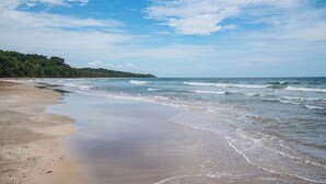 A beach with a sandy shore and gentle waves, a clear sky, and a forested hill in the background. - Villa Oasis (Cahuita)