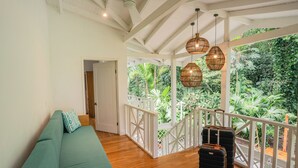 A room with a green sofa, two suitcases, wicker pendant lights, and a view of lush greenery through a white balcony railing. - Villa Oasis (Cahuita)