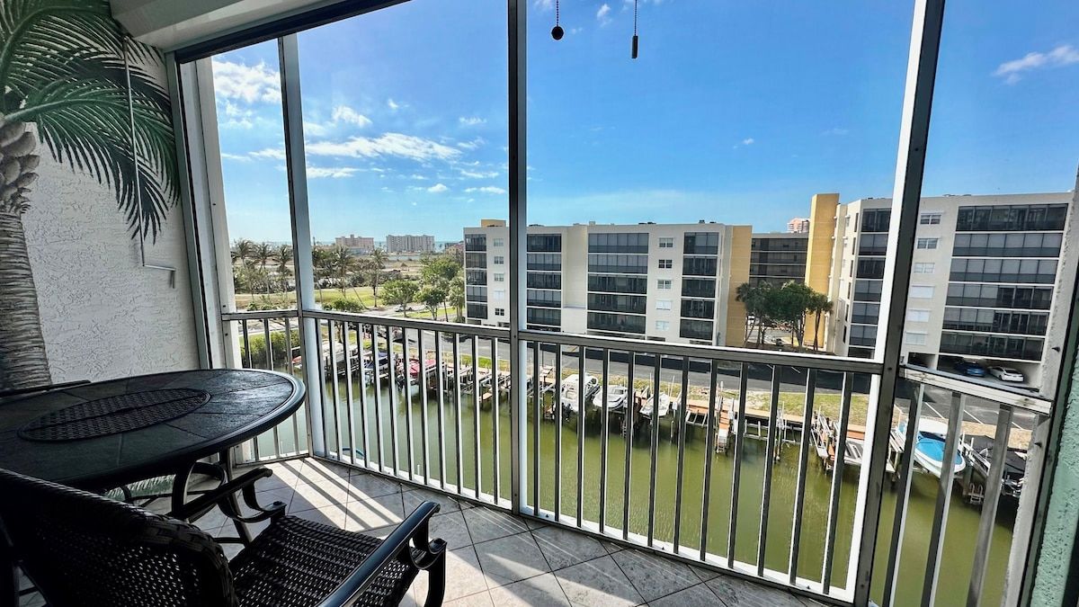 A balcony with a table and chairs, overlooking a marina with boats and buildings in the background.