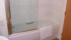 Kylpyhuoneessa on lasiseinällä varustettu suihku, valkoinen kylpyamme ja valkoinen pyyhe, joka on ripustettu ammeen reunalle.