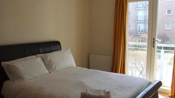 Hotellihuone, jossa on sänky, patteri ja ikkunasta avautuva näkymä toiseen rakennukseen, ja ikkunassa on verhot.