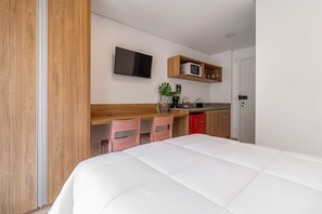 A modern hotel room with a bed, a desk with a computer, a microwave, and a red refrigerator.