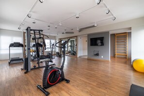 Uma academia ampla com diversos equipamentos de exercício, piso de madeira e uma grande janela que proporciona iluminação natural.