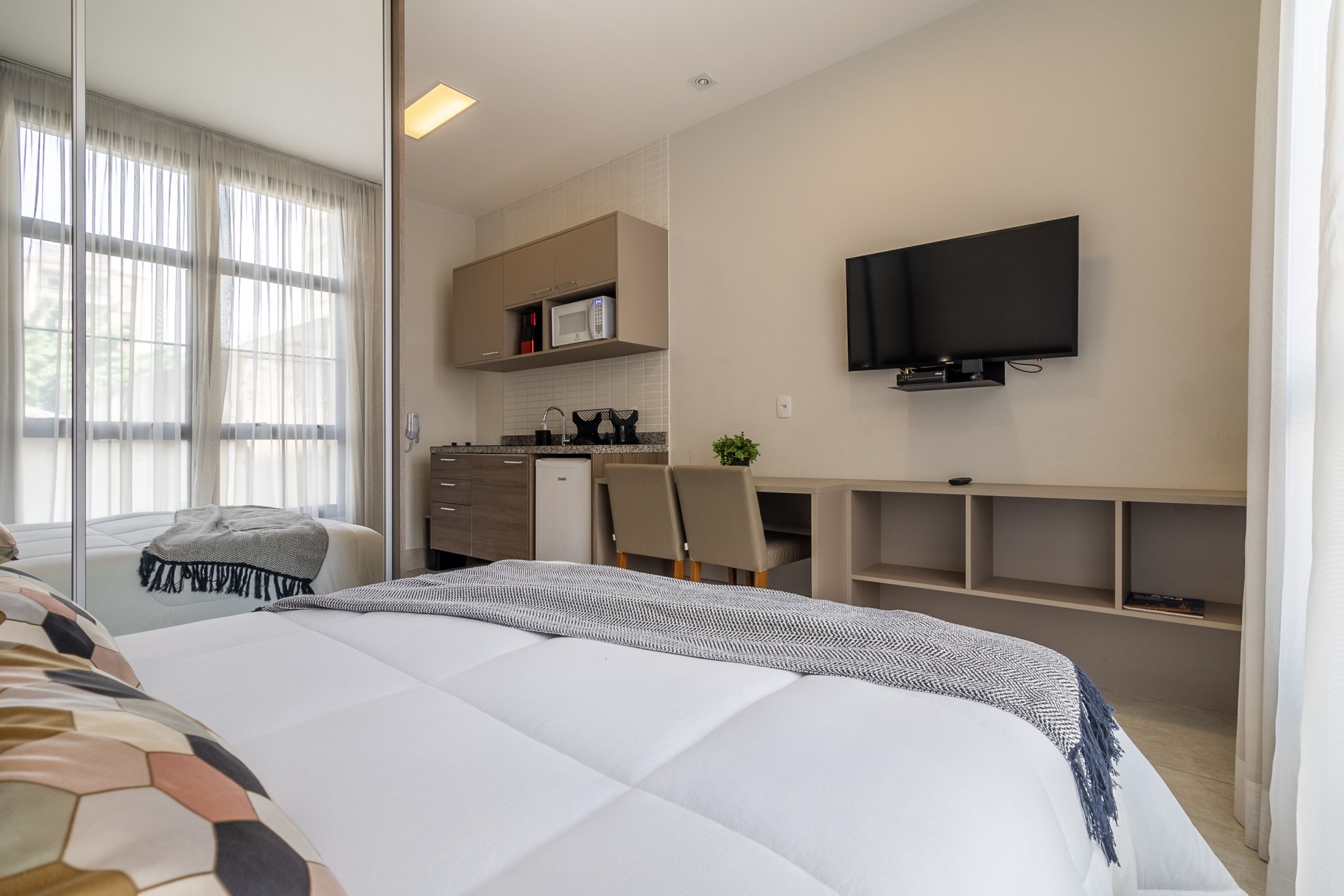 Ein modernes Hotelzimmer mit einem großen Bett, einem Schreibtisch mit Stuhl, einem Fernseher und einer Küchenzeile.