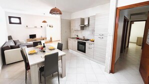 A modern kitchen with white cabinetry, a dining area with a table and chairs, and a living area with a sofa and TV. - Casa Amodei 2 a Pochi Passi dal Mare (San Vito Lo Capo)
