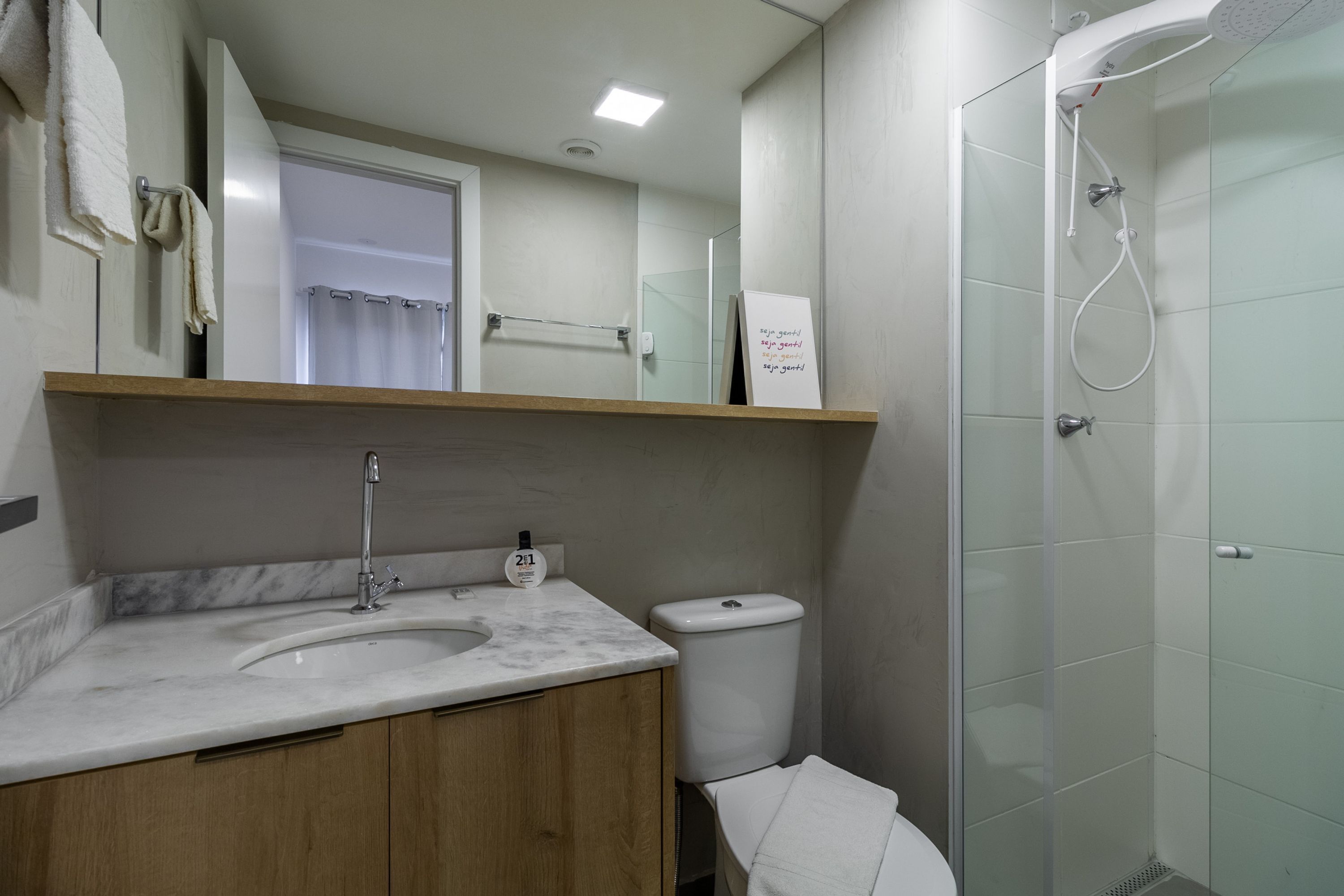 Ein Badezimmer mit Marmorwaschbecken, Holzwaschtisch, Toilette, Duschkabine aus Glas und einem weißen Handtuch auf dem Boden.