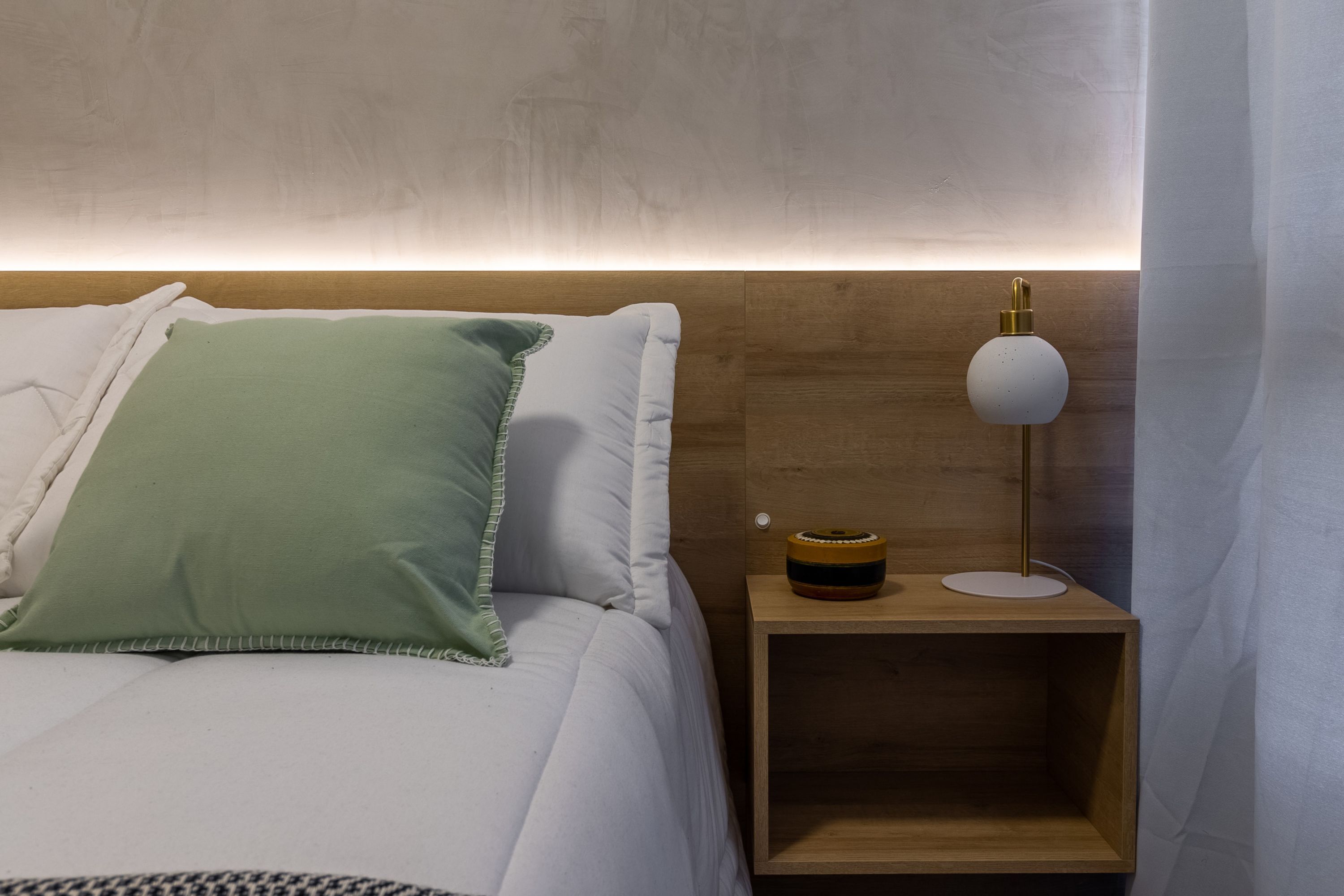 Ein ordentlich bezogenes Bett mit weißen und grünen Kissen, ein Nachttisch mit Lampe sowie ein Holz-Kopfteil.