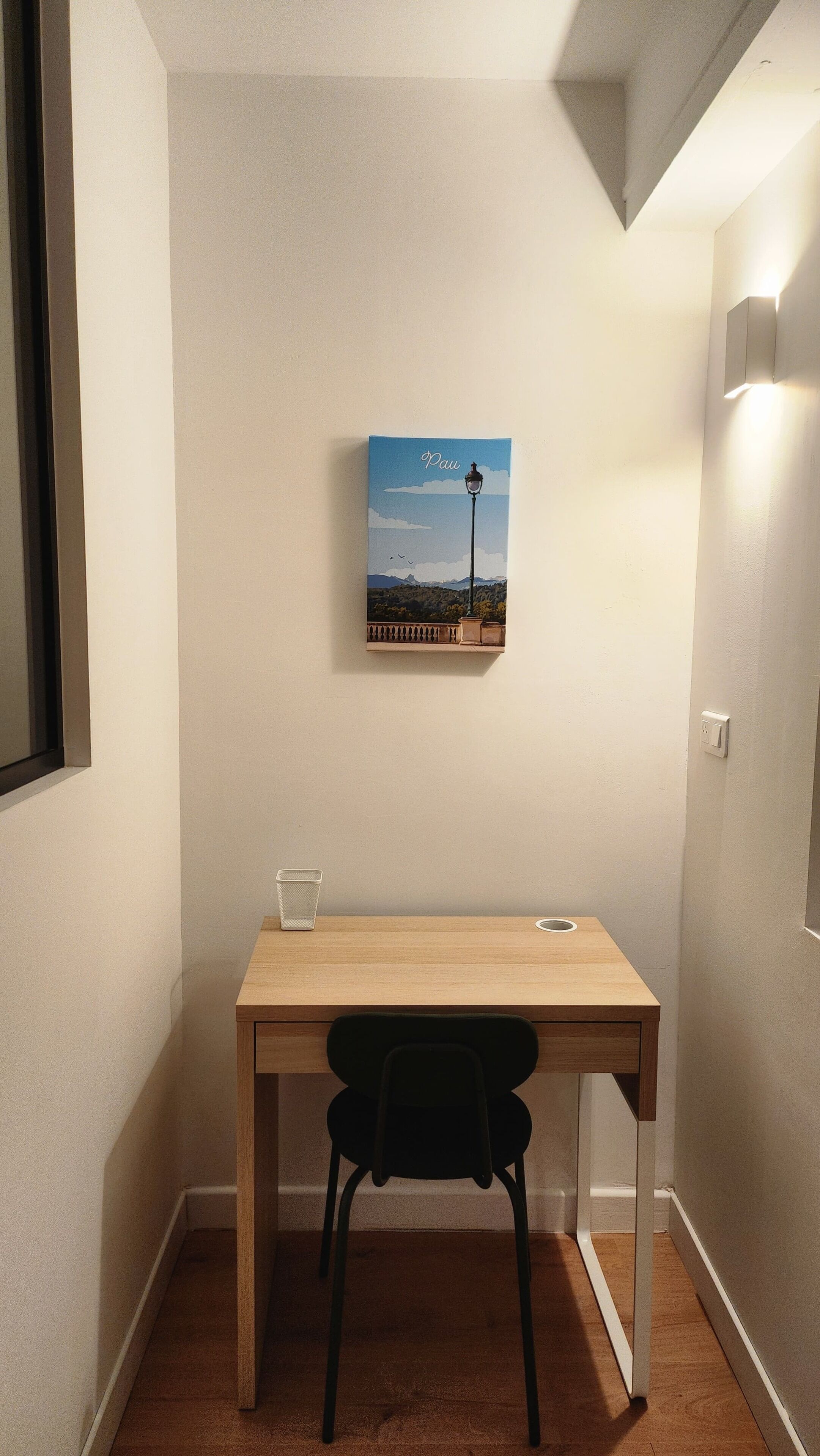 Un espacio de trabajo pequeño y ordenado, con un escritorio, una silla, un cuadro en la pared y un interruptor de luz.