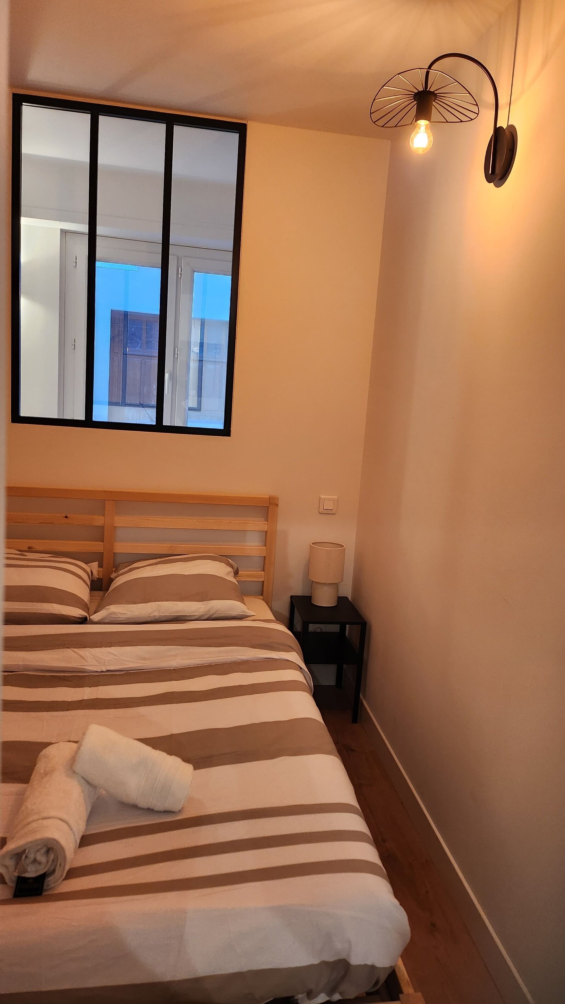 Een slaapkamer met een houten bed, gestreept beddengoed, een raam met uitzicht, een bedlamp en een wandlamp.
