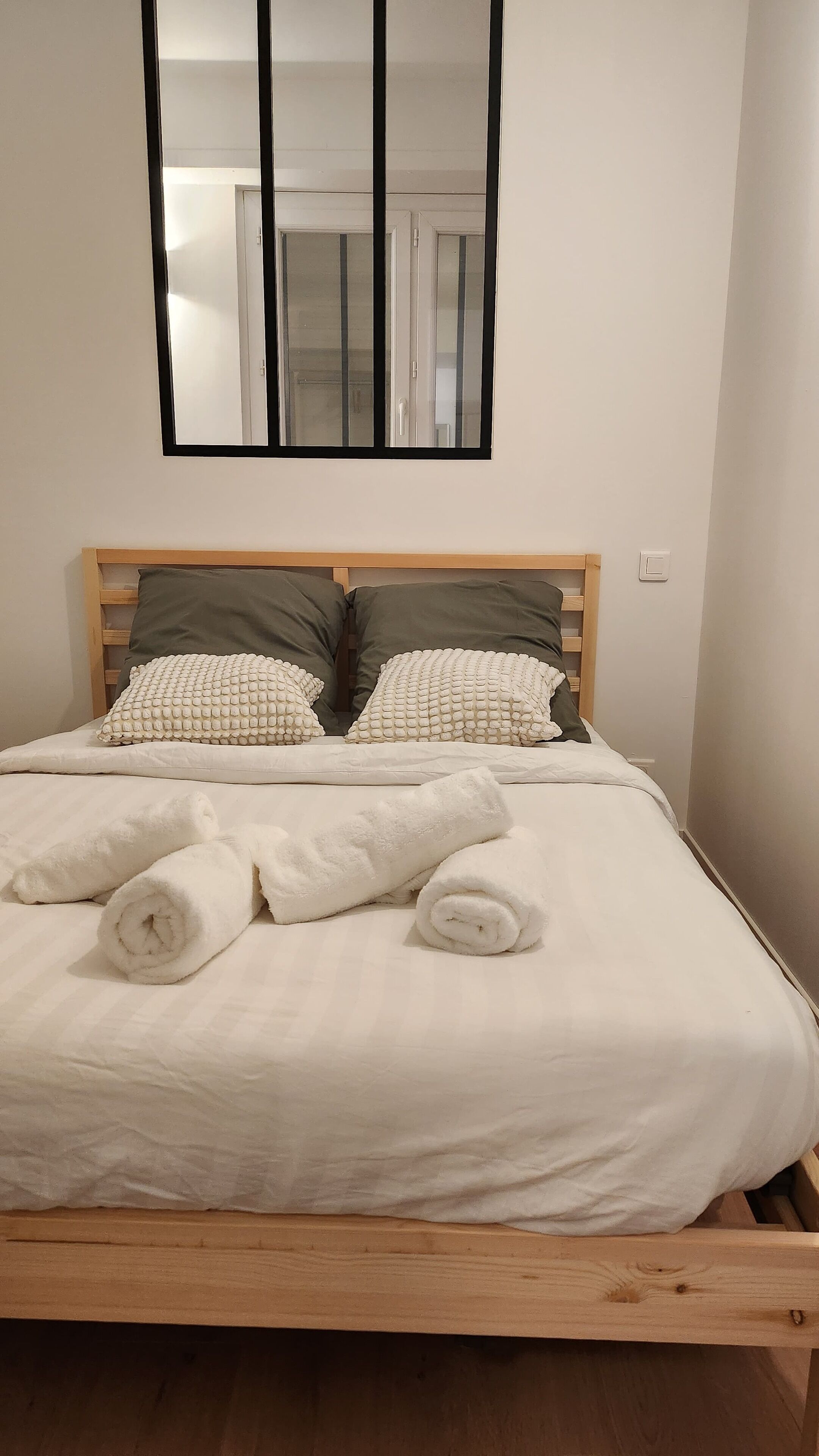 Ein ordentlich gemachtes Bett mit weißen Leinentüchern und aufgerollten Handtüchern, ein Holz-Kopfteil und ein Spiegel darüber.