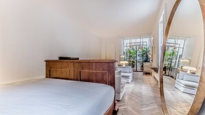 Une chambre avec un grand lit, une commode en bois et un miroir. On y trouve également un coin salon avec un canapé et des fauteuils.