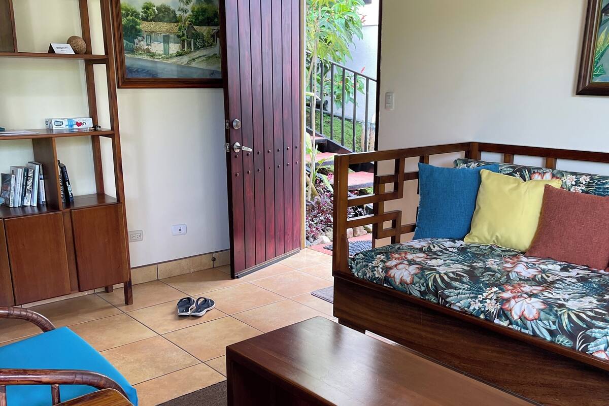 Una habitación con un sofá de madera, una mesa de centro de madera, una silla azul, una estantería y una puerta que da al exterior.