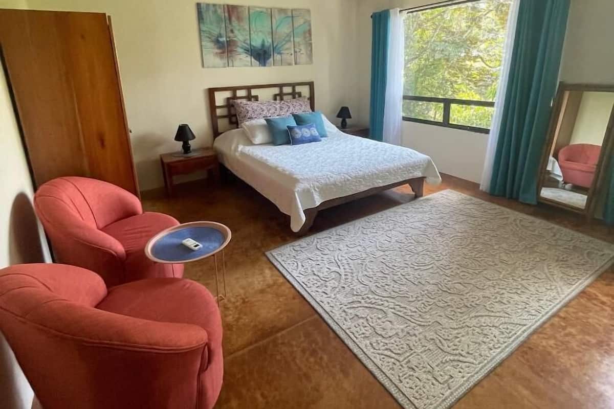 Un dormitorio con una cama, un ventilador de techo, una mesita de noche, un sillón rojo, un otoman azul y un ventanal con cortinas.