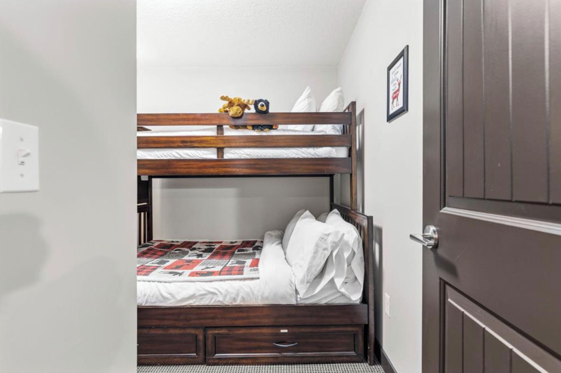 A bunk bed with a quilt, pillows, and a small stuffed animal on the top bunk. There is a door to the right and a light switch on the wall.