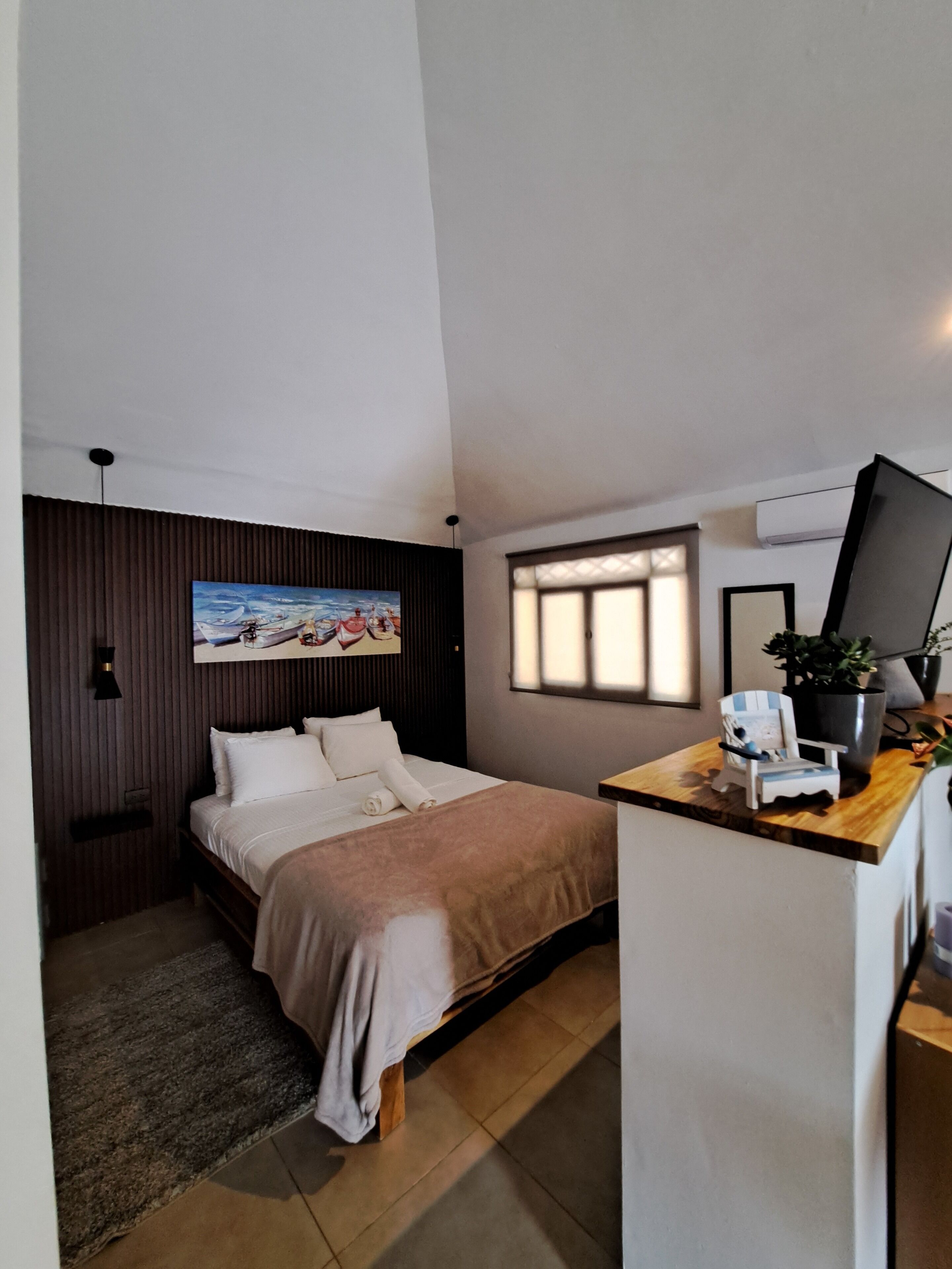A compact hotel room with a bed, a small desk, a potted plant, and a painting on the wall.