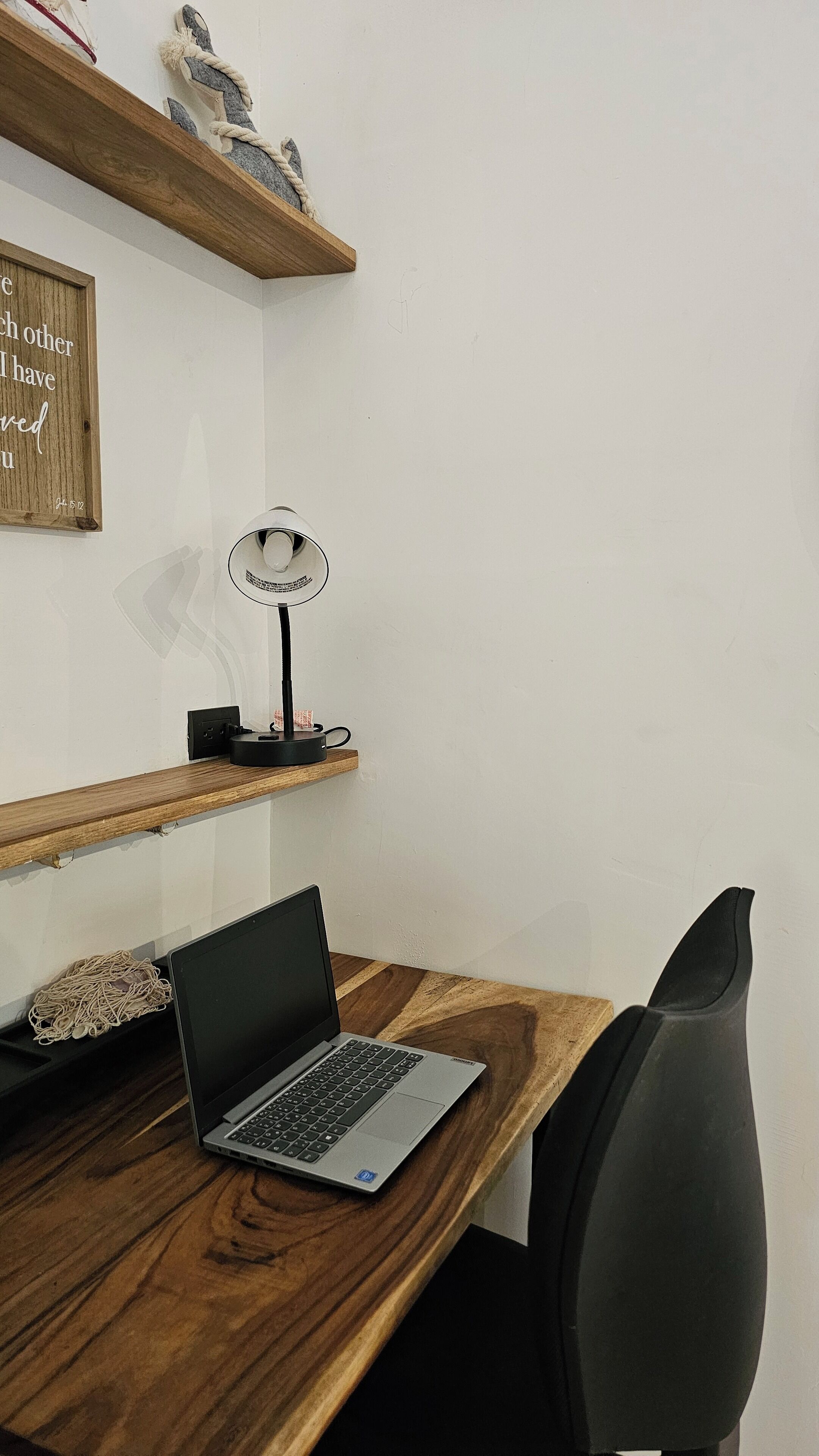 Un bureau en bois avec un ordinateur portable, une lampe de bureau et une chaise. Au-dessus du bureau, on trouve des étagères en bois ornées d’objets décoratifs.