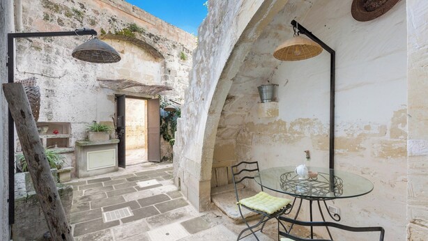 A stone courtyard with a glass table and chairs, a hanging lamp, and a rustic door.