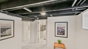 Un couloir aux murs blancs, orné de tableaux représentant des paysages urbains, avec un tabouret en bois sur lequel est posée une plante, et une porte donnant sur une autre pièce.