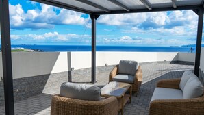 An outdoor patio with wicker furniture, a view of the sea, and a clear sky. - Ozeanica Heights by LovelyStay (Gaula)