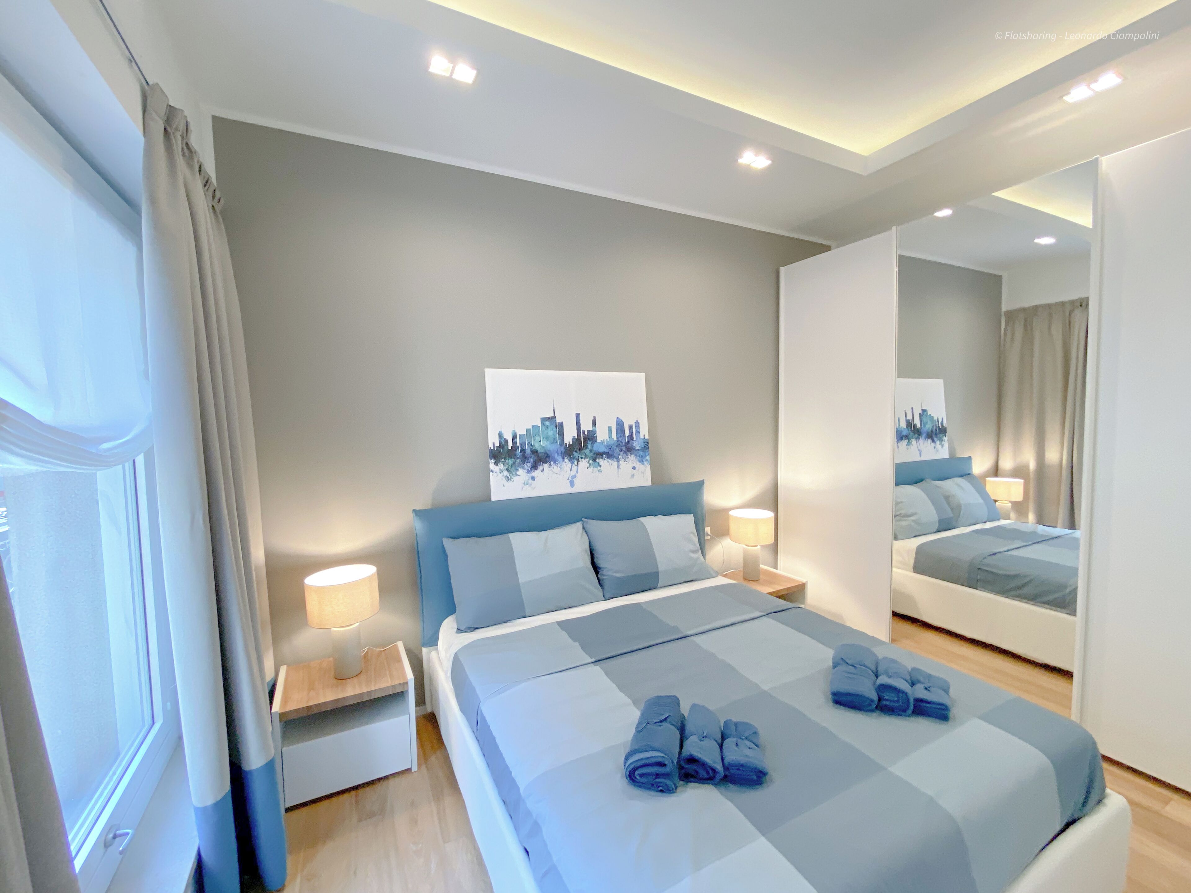 A modern hotel room with a large bed, a nightstand, a mirror, and a cityscape artwork on the wall.