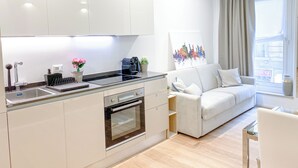Eine moderne Küche mit weißen Schränken, einem eingebauten Ofen und einer Kaffeemaschine. Im Wohnbereich befindet sich eine weiße Couch und ein Fenster mit Vorhängen.