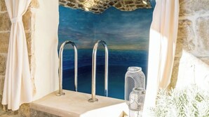 A stone bathtub with two faucets, a decorative glass container, and a view of the sea through a textured stone wall. - Residence Villa Mima (Cavtat)