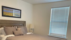 Quarto com cama, ventoinha no teto, janela com persianas, mesa de cabeceira, abajur e quadro na parede.