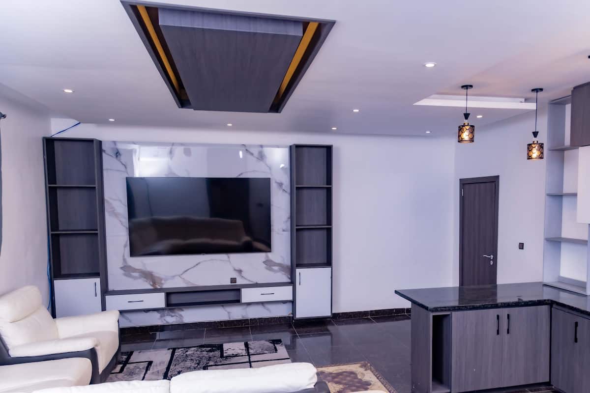 A modern living room with a large flat-screen TV, built-in shelving, a sofa, and a kitchen area with dark cabinets and a marble countertop.
