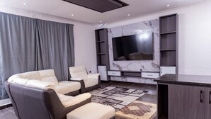 A modern living room with a flat-screen TV, grey and white leather sofas, a marble-patterned wall, and built-in shelving. - Pinorich Villa (Buea)
