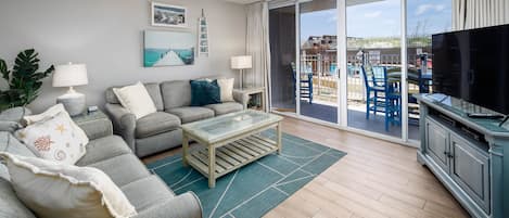 A living room with a sofa, coffee table, and TV, a balcony with chairs, and a view of the beach.