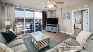 Sala de estar com mesa de centro de vidro, sofás cinzas, televisão e vista para uma piscina e área de estar externa.