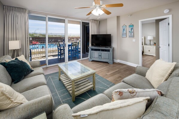 A living room with a glass coffee table, grey sofas, a TV, and a view of a pool and outdoor seating.