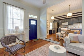 Un salon spacieux avec une porte bleue, un parquet en bois, un fauteuil à motifs, un canapé blanc et un coin cuisine équipé d’appareils électroménagers modernes.