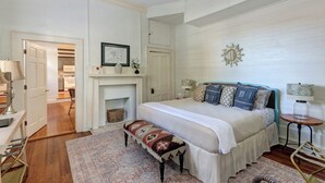 Une chambre à coucher comprenant un lit, des tables de chevet, un banc, une cheminée et donnant sur la cuisine.