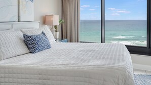 A bedroom with a large bed, a nightstand with a lamp, a potted plant, and a view of the ocean through large windows. - VillaSolare-Sandestin: 9th-Floor Gulf-Front+Views (Miramar Beach)